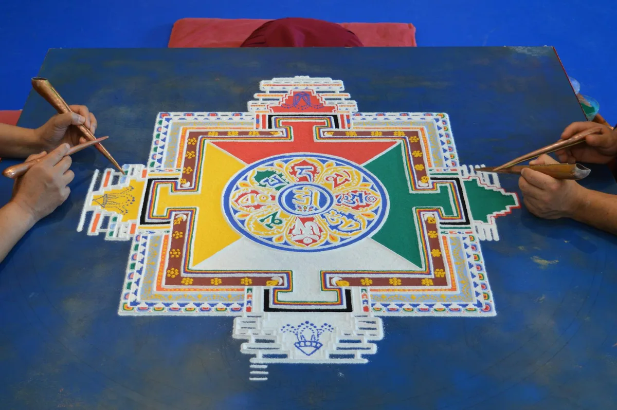 Nahaufnahme von Händen bei der Erstellung eines farbenfrohen tibetischen Sandmandalas mit Präzisionswerkzeugen auf blauem Hintergrund