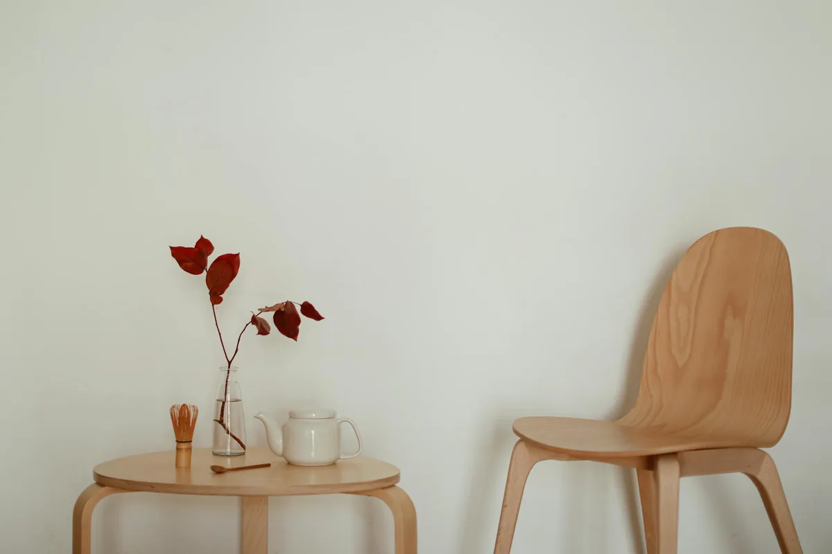 Interno minimalista con tavolo, foglie rosse in vaso, teiera e sedia di legno su parete neutra