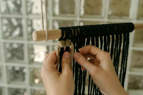 Hands weaving a macrame wall hanging using black and beige yarn. Arts and crafts in focus.