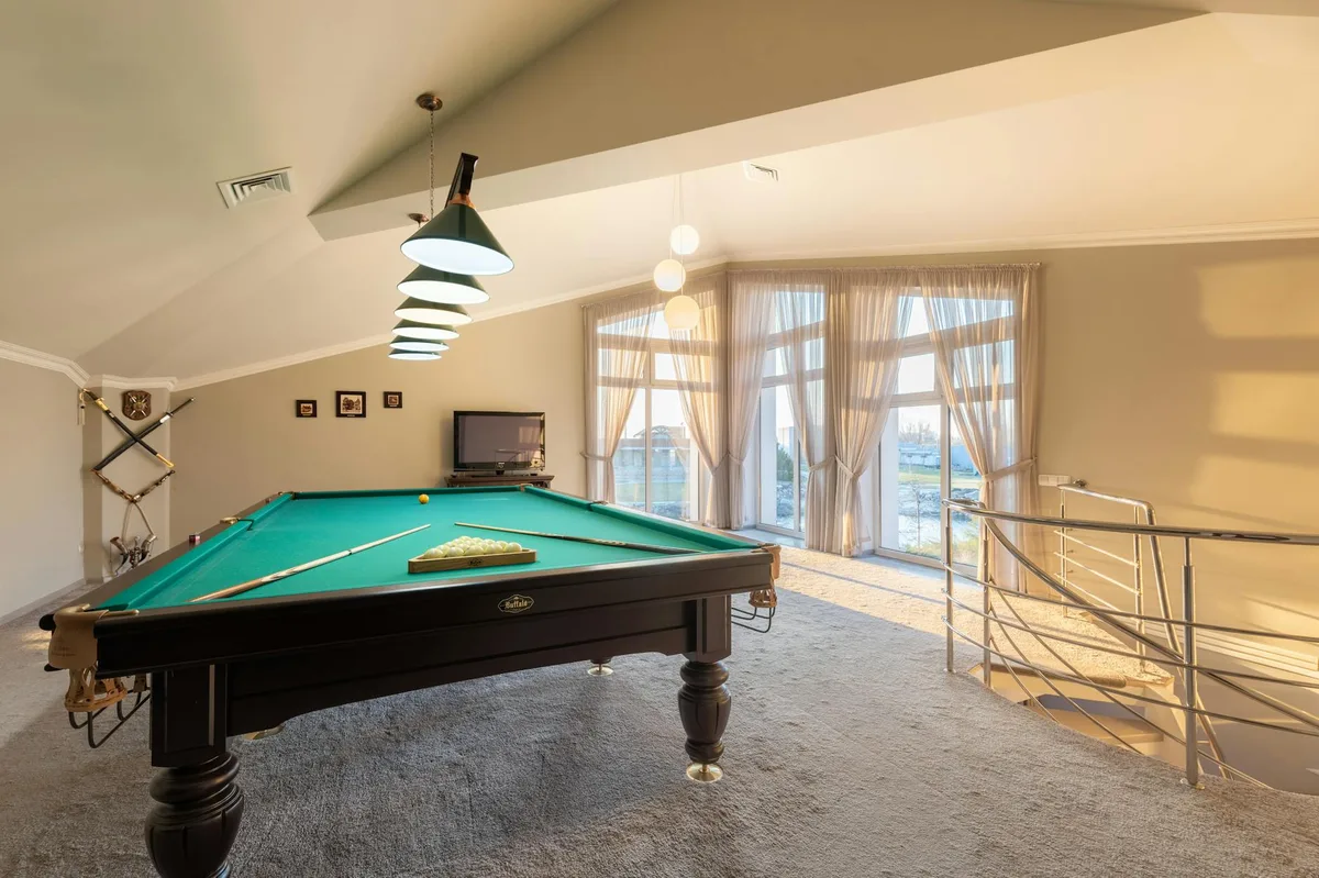 Table de billard en bois avec queues sur tapis dans une pièce spacieuse avec fenêtres et escalier à rampe métallique