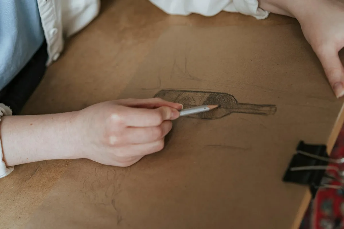 Person skizziert eine Weinflasche auf Papier mit einem Bleistift im kreativen Prozess