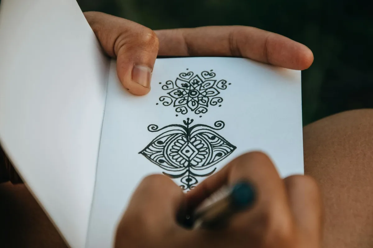 Crop anonymous talented person creating difficult mehendi sketch in album with white sheets