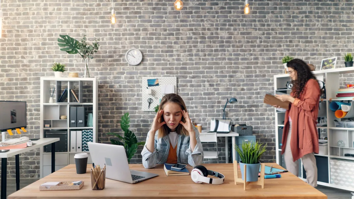Two women in a stylish office setting, one focused at a desk, another reviewing notes.