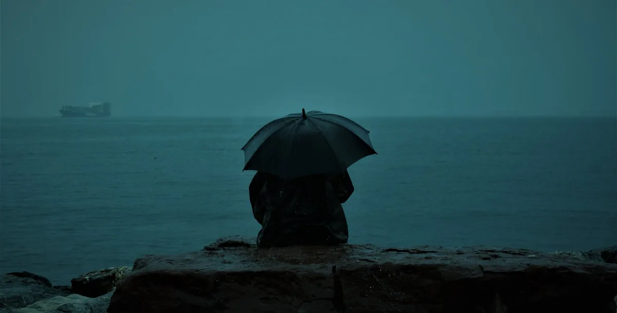 孤独身影持伞坐于雨中伊兹密尔海边，唤起孤独与内省的图像。