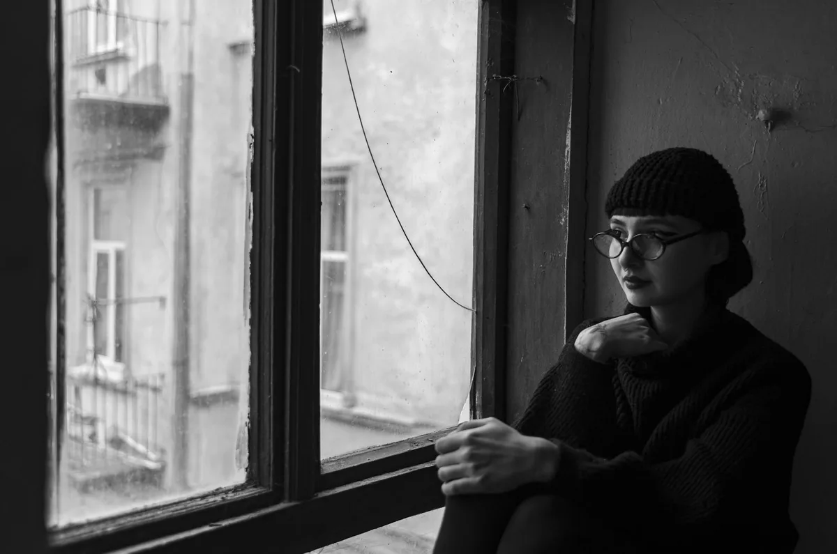 Ritratto in bianco e nero di donna con berretto e occhiali seduta vicino a finestra