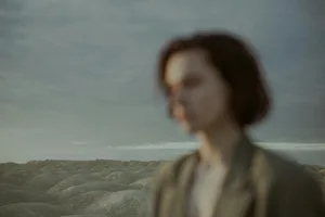 Blurred portrait of a woman in front of a serene, rolling landscape with a muted sky.