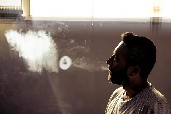 Side profile of a man blowing smoke indoors with dramatic lighting.