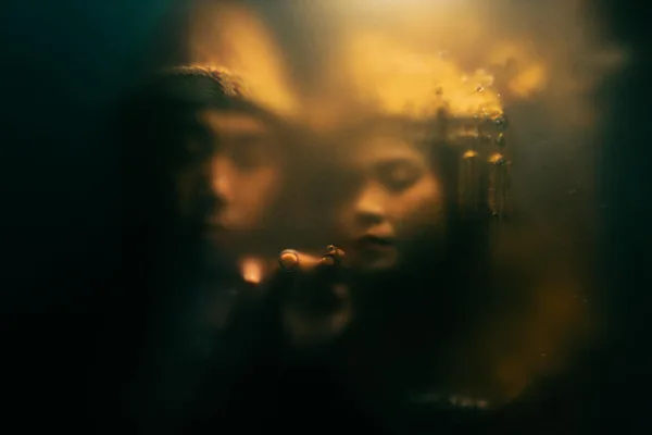 Two people create an enigmatic scene behind a steamy glass with warm lighting.