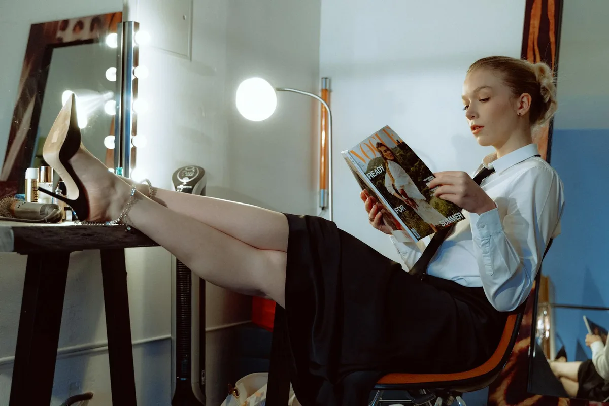 Stilvolle Frau in der Umkleide liest Modemagazin mit entspannten Beinen auf dem Schminktisch.