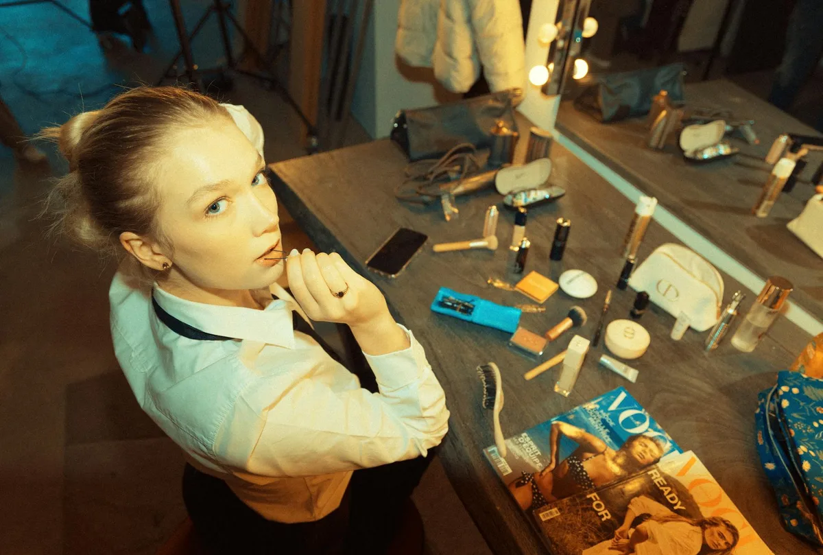 A young woman preparing in a dressing room with makeup products and magazines around.