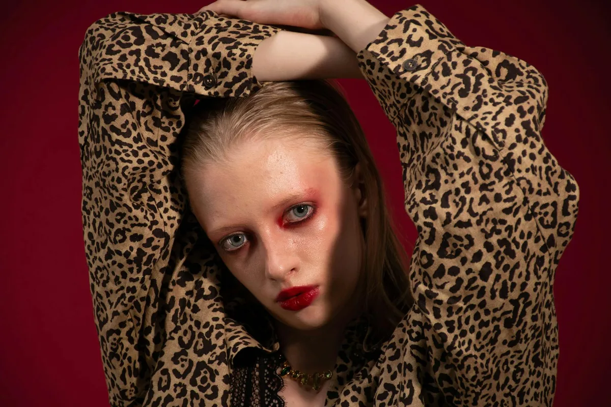 Modeporträt einer Frau in Leopardenbluse mit erhobenen Armen und mutigem Make-up auf rotem Hintergrund