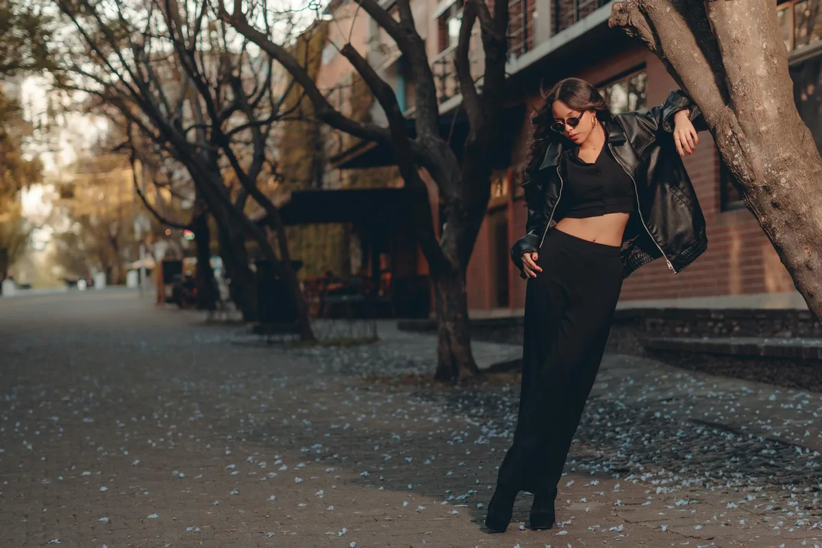 Модная женщина в чёрной кожаной куртке позирует на городской улице с деревьями