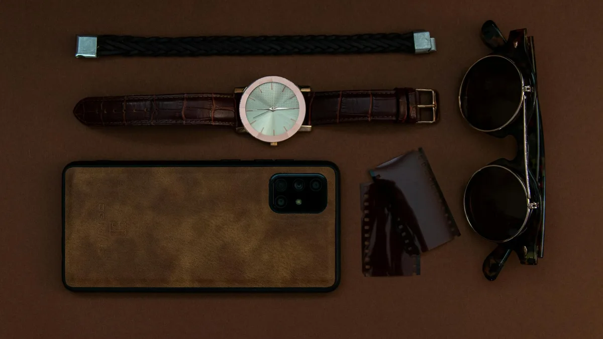 Minimalistisches Flatlay mit Uhr, Sonnenbrille, Smartphone und Lederriemen auf brauner Oberfläche