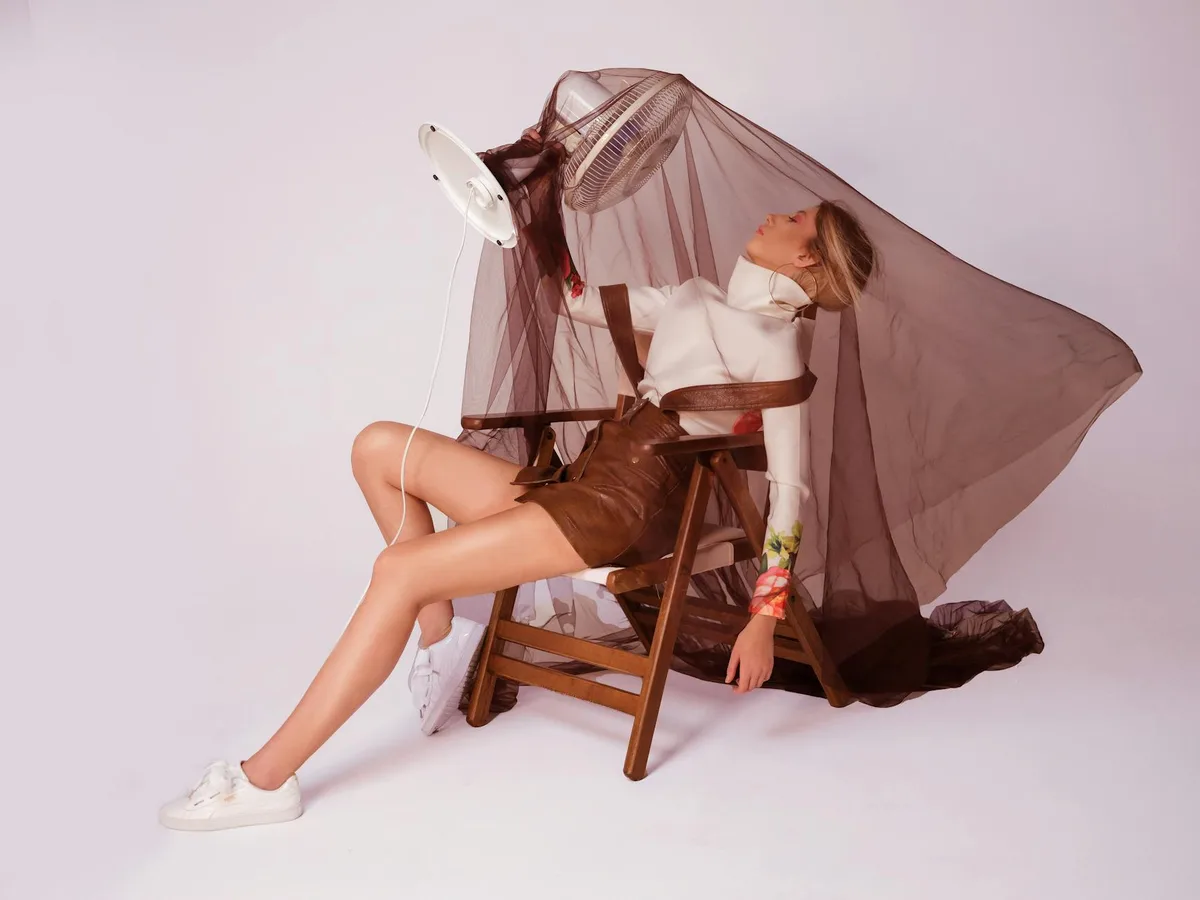 Mannequin assis sur chaise en bois sous tissu transparent avec ventilateur, vue de côté.