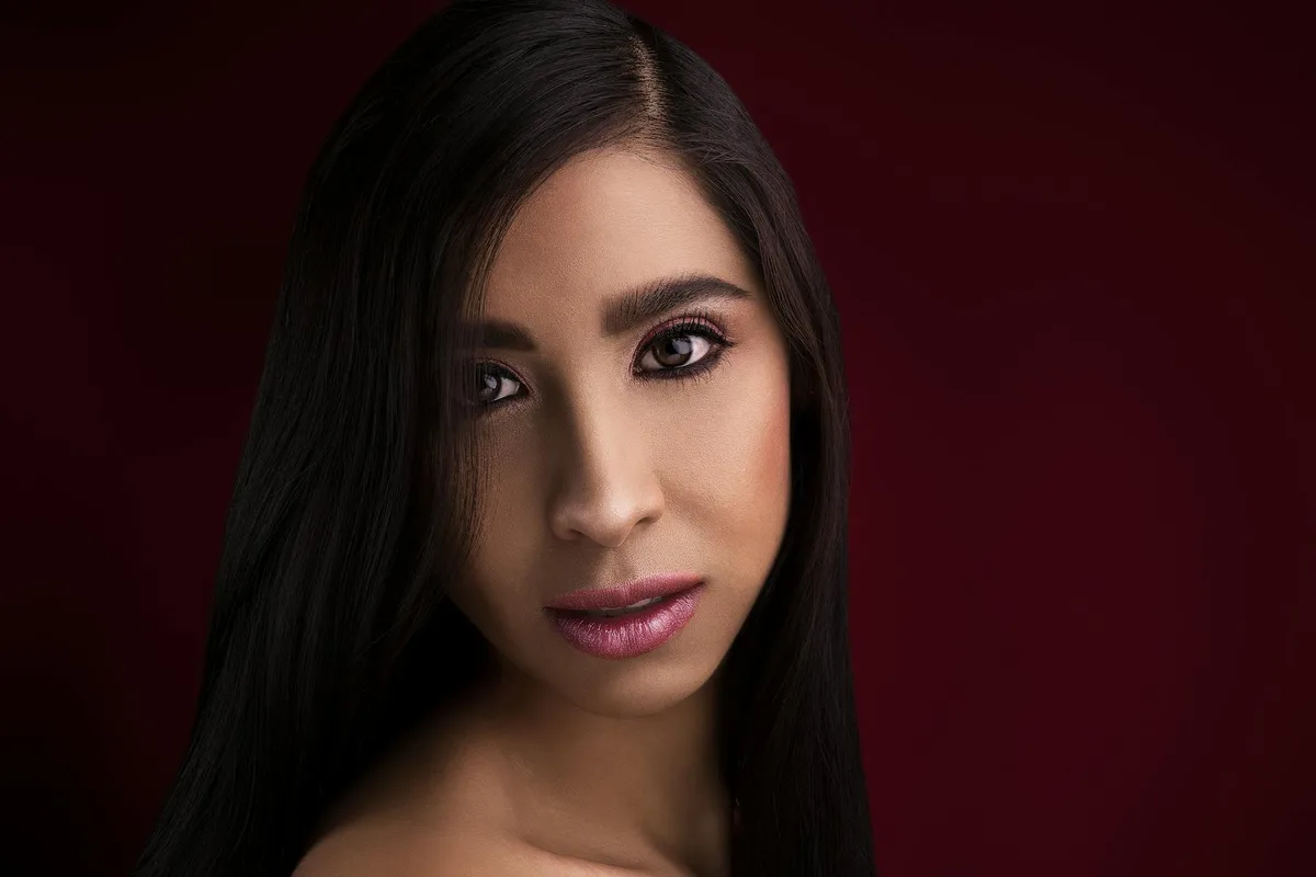 Studio portrait of a woman with long dark hair and pink lipstick against a dark background.