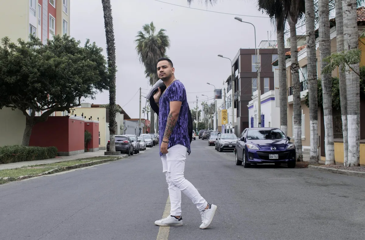 Uomo elegante in jeans bianchi e camicia viola posa in strada cittadina con auto