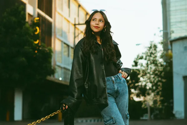 Stilvolle Frau in Lederjacke und Jeans in der Stadtsilhouette bei Dämmerung