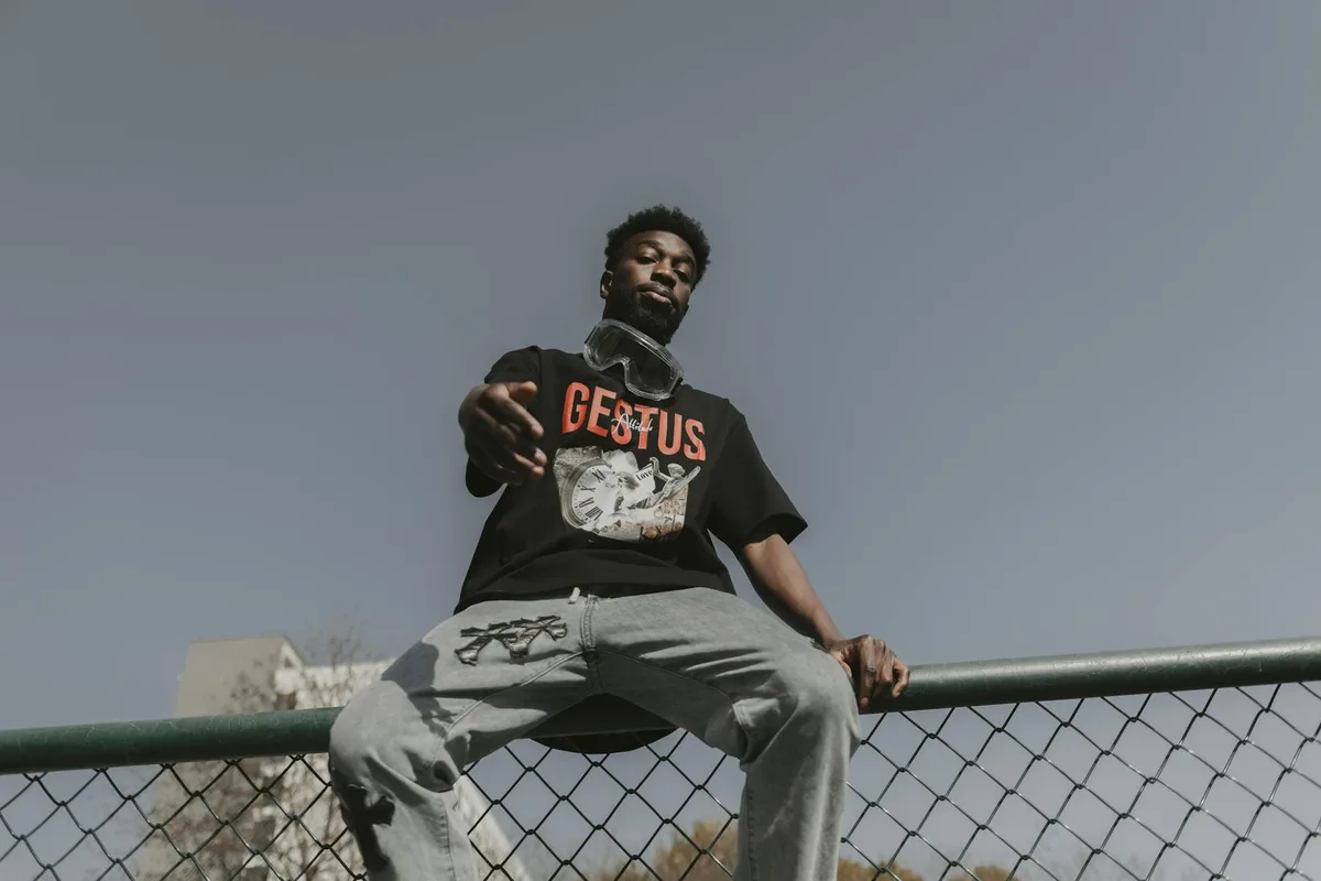 Jovem estiloso sentado em cerca ao ar livre com pose confiante.