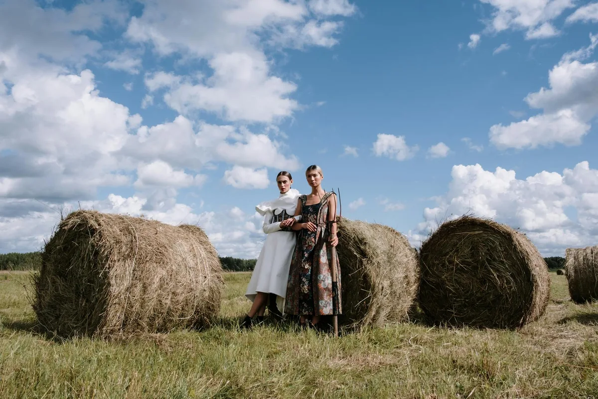 Zwei Models in stylischen Outfits posieren zwischen großen Strohballen auf einem ländlichen Feld