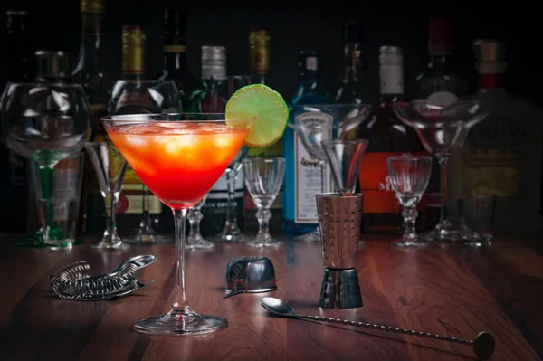 Lebhafter Cocktail mit Limettengarnitur umgeben von Barwerkzeugen und Flaschen