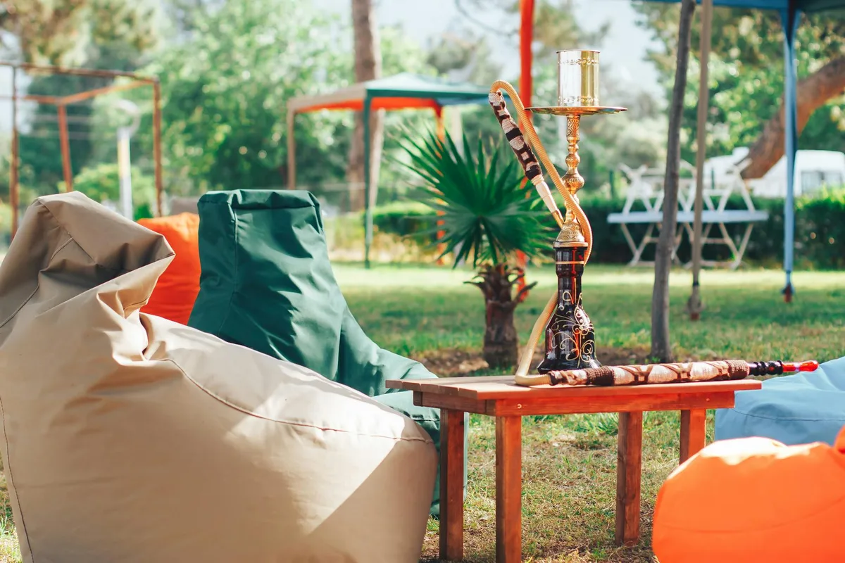 Pouf morbidi su prato vicino a tavolo di legno con narghilè in parco verde di giorno