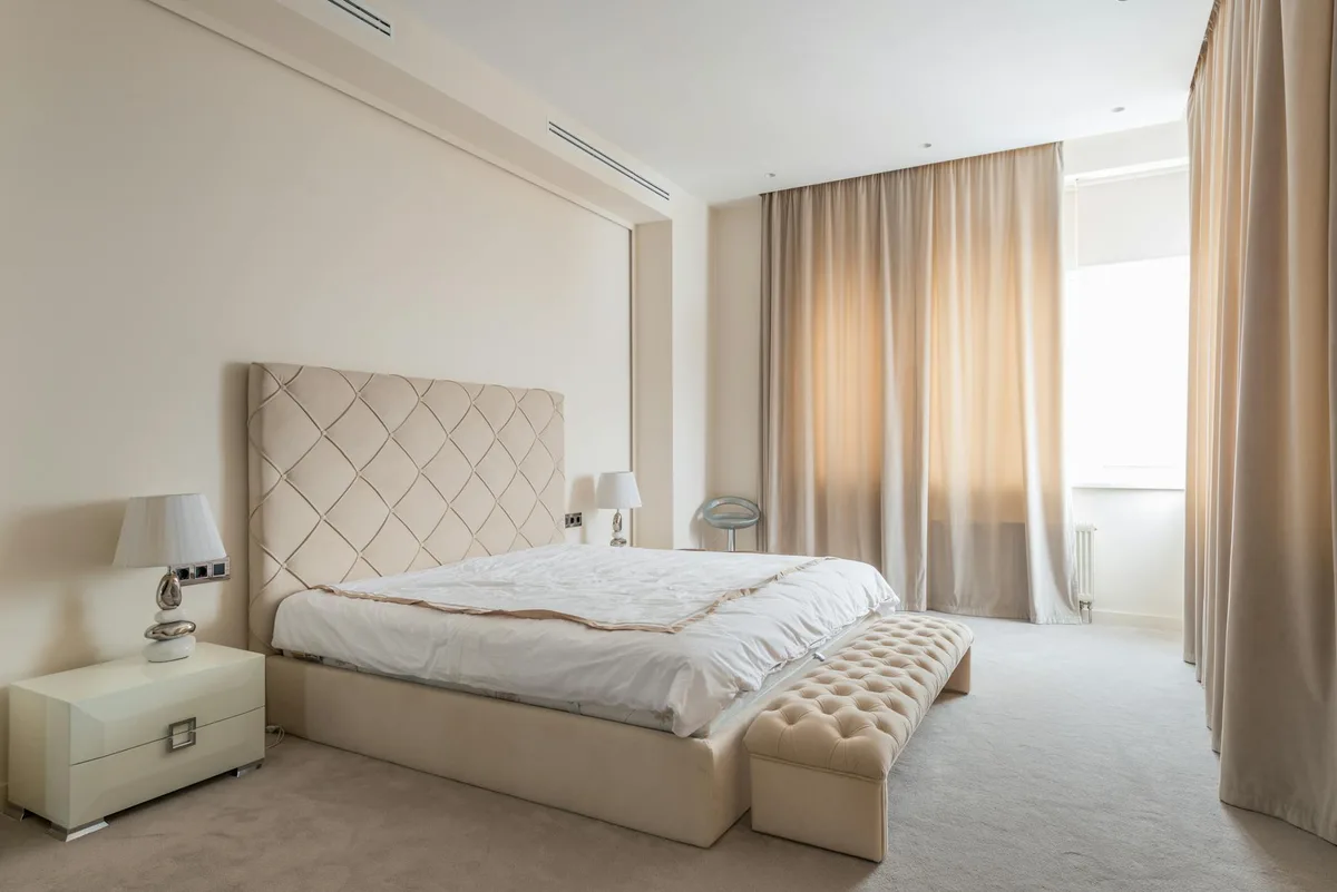 Comodini con lampade eleganti accanto a letto con testiera beige in camera da letto spaziosa con panca e tende