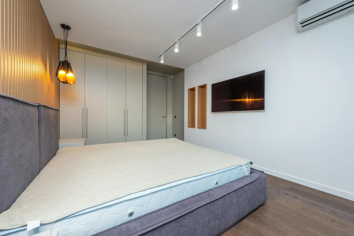 Camera da letto moderna spaziosa con letto grigio e TV a parete con illuminazione ambientale