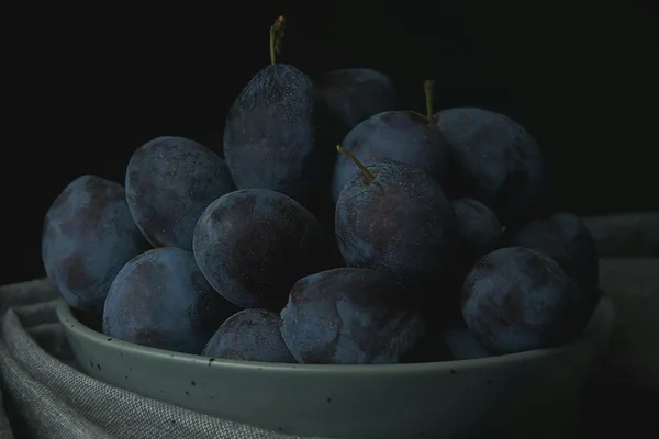 A close-up photo of a bowl filled with fresh, ripe purple plums on a black background.