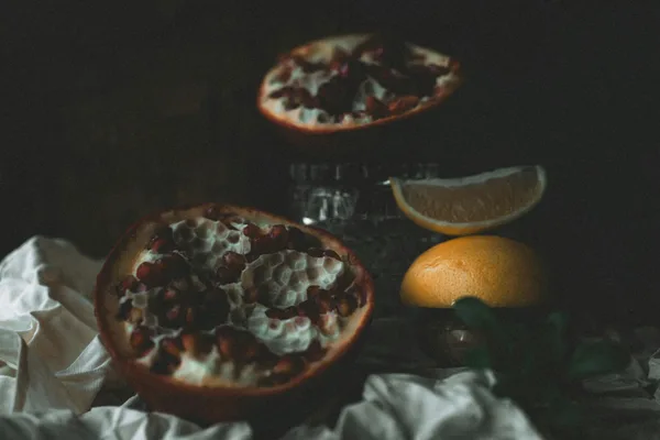 A dark, moody still life featuring pomegranate halves and citrus on fabric.