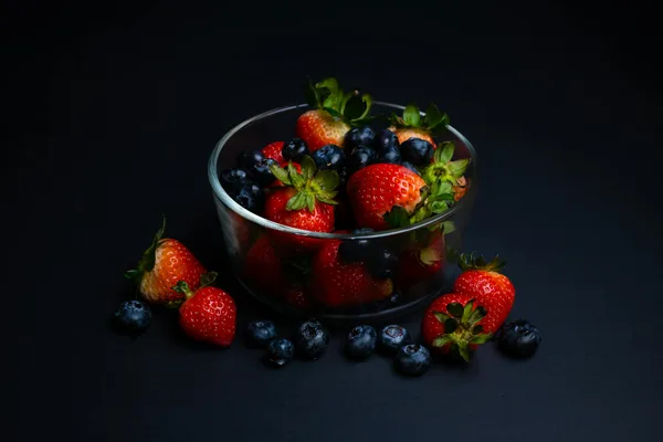 A glass bowl filled with fresh strawberries and blueberries against a dark background.
