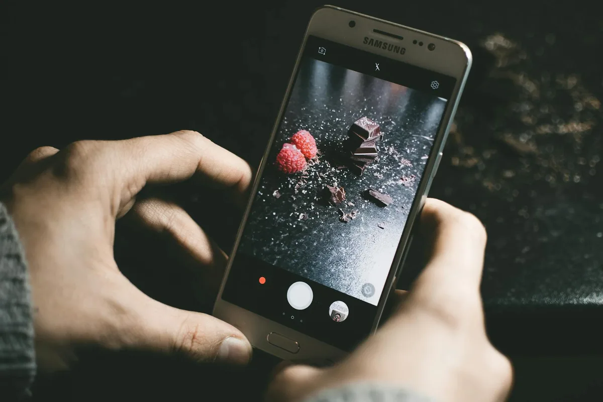 Nahaufnahme eines Smartphones, das Schokoladenstücke und Himbeeren auf einer dunklen Oberfläche fotografiert.