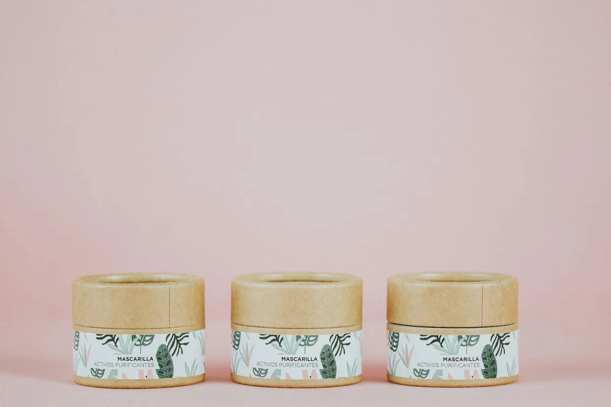 Tre barattoli skincare ecologici con design botanico su sfondo rosa, packaging sostenibile