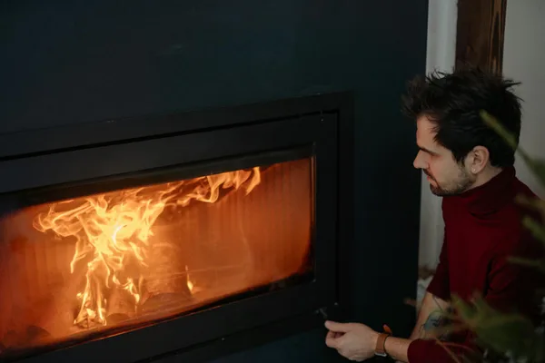 Man in cozy red sweater enjoying warmth from modern fireplace indoors.