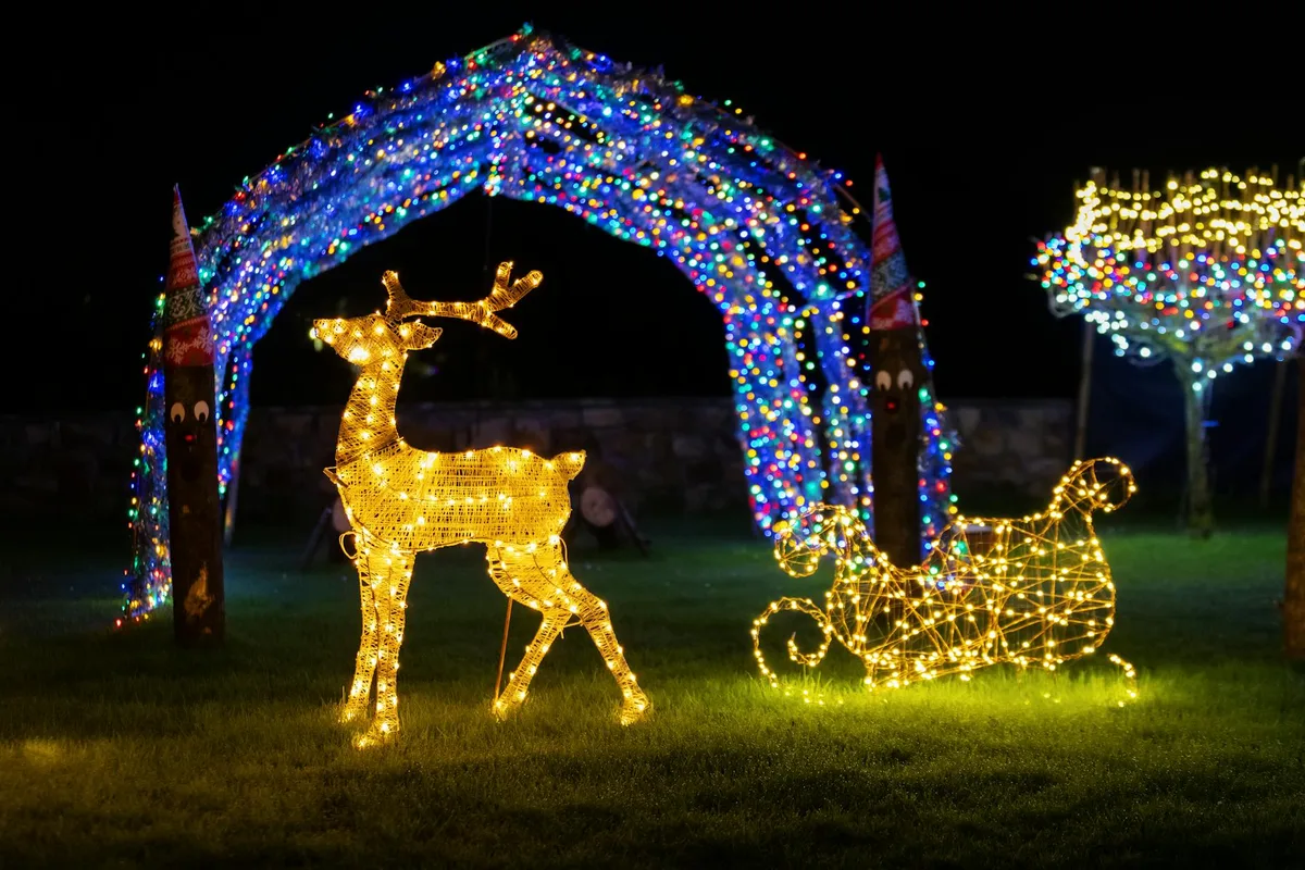 Яркая уличная инсталляция с подсвеченными оленями и санями под разноцветной аркой для праздничного ночного вида