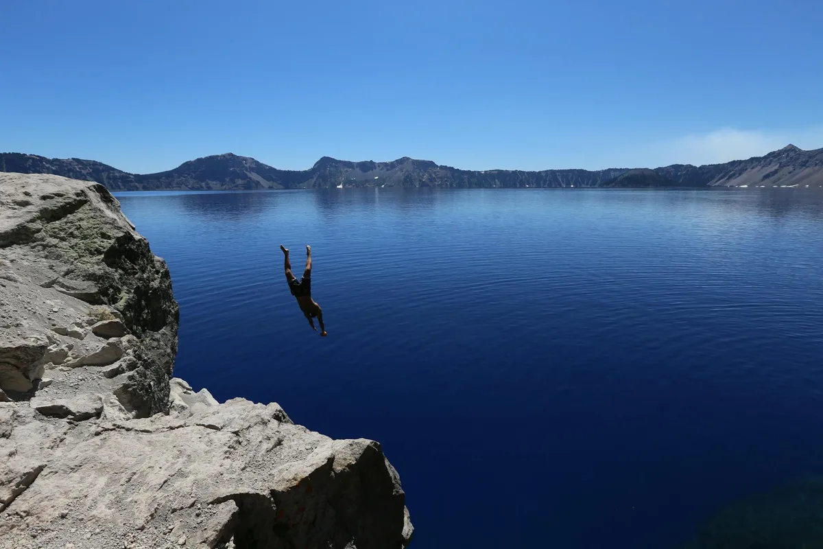 Persona che si tuffa da una scogliera rocciosa nel Crater Lake, acque blu sotto un cielo luminoso.