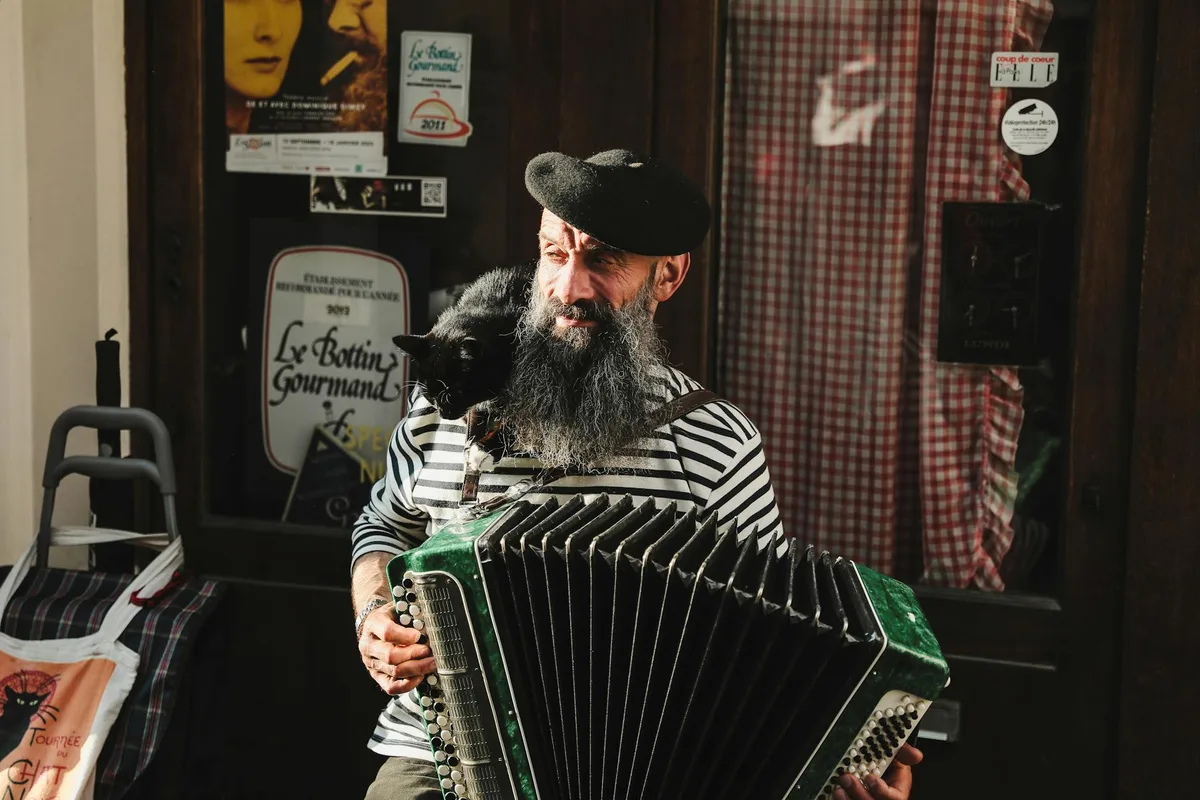 Bearded street musician playing accordion with a black cat perched on his shoulder.