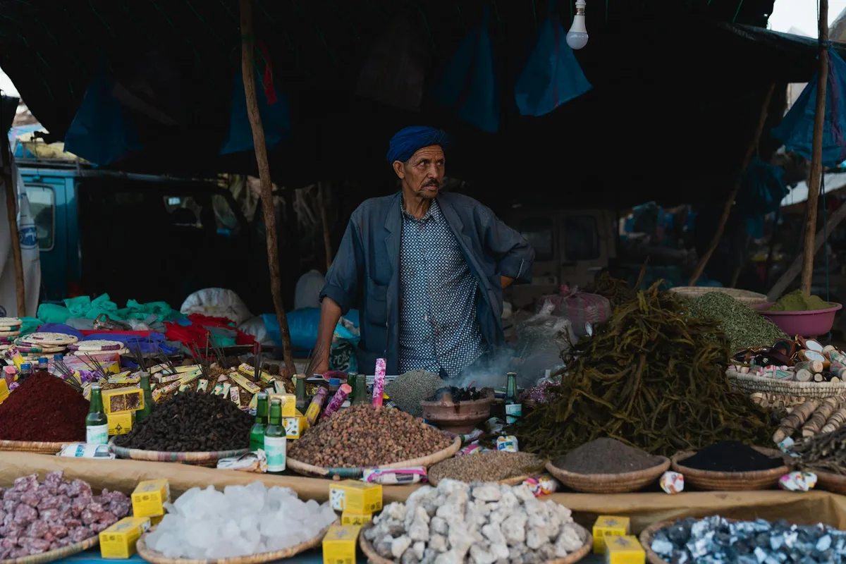 Homme d'âge mûr en tenue traditionnelle vendant des épices sur un marché animé.