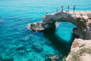 Tourists enjoying a sunny day cliff diving at a stunning natural rock arch over clear blue waters.