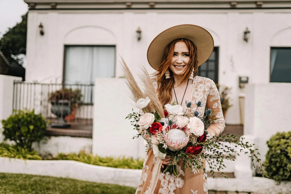 Sposa boho sorridente con bouquet davanti a casa rustica per temi wedding
