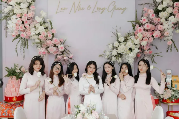 A bridal party of young women in traditional pink Ao Dai dresses holding flowers at a wedding celebration.