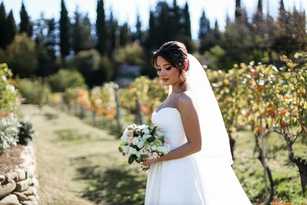 Braut in elegantem Kleid hält in einem malerischen Weinberg einen Blumenstrauß