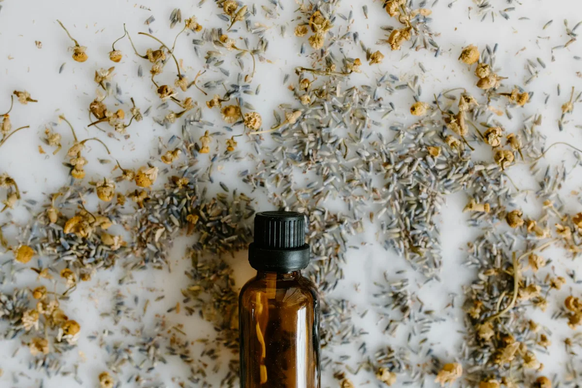 Primo piano di bottiglia di olio essenziale con lavanda e camomilla essiccate su superficie bianca.