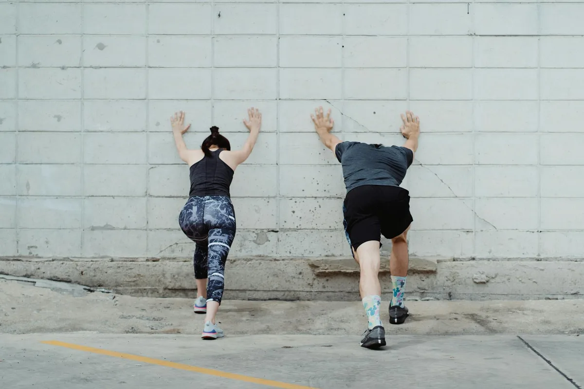 Casal em roupas esportivas se alonga contra parede ao ar livre