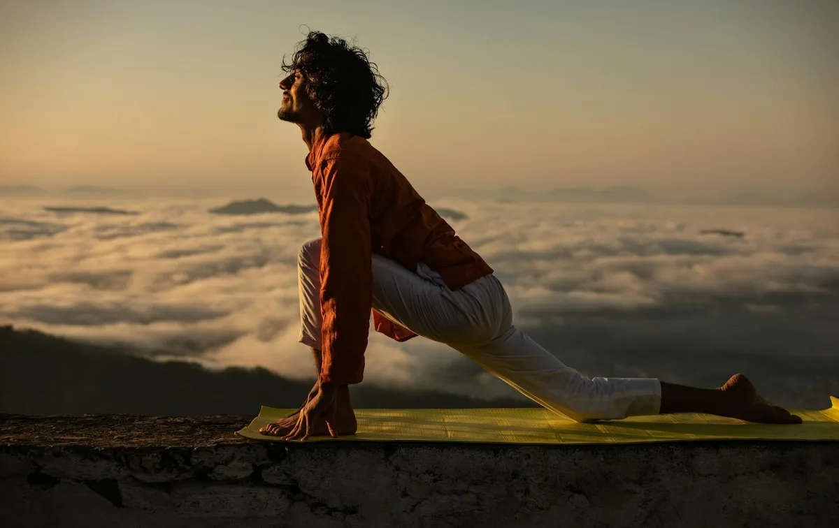 Ein Mann führt eine Yoga-Pose auf einer Matte mit atemberaubendem Sonnenaufgang über den Wolken aus.
