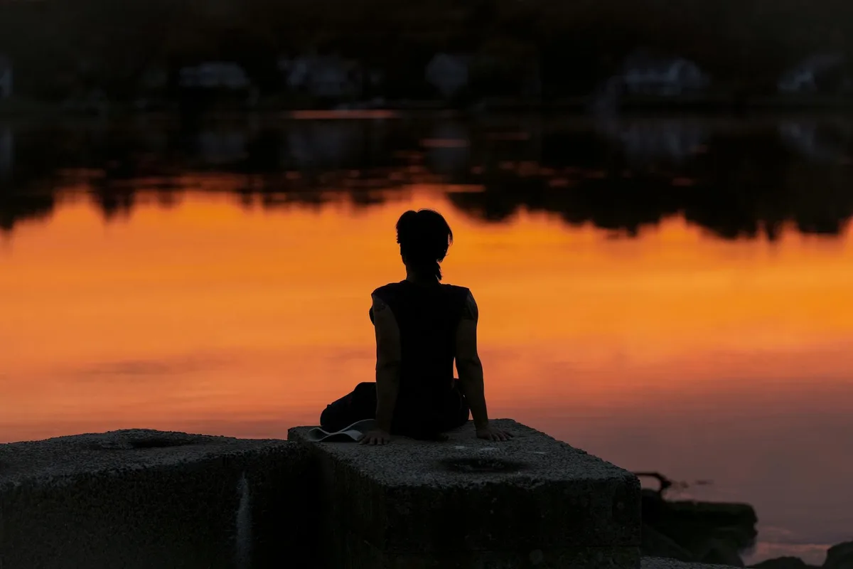 Persona medita all'alba in riva a un lago calmo, silhouette serena.